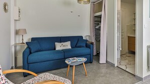 Una sala de estar con un sofá azul, una mesita con libros, una lámpara y una puerta que da a un baño.