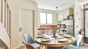 Un comedor con una mesa redonda dispuesta para cuatro, una cocina al fondo y una escalera que sube a otro nivel.