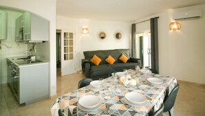A dining area with a table set for four, a sofa with cushions, a kitchen area, and a wall-mounted air conditioning unit. - FLH Armação Pêra Beachside Bliss (Armação de Pêra)