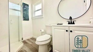 A bathroom with a glass shower enclosure, a toilet, a sink with a mirror, and a logo for Timber Valley Vacation Rentals. - Cozy 1BR Entire Unit in W. Plains, nice location! (West Plains)