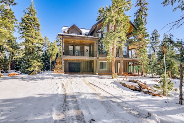 A two-story log cabin with a garage, surrounded by snow and trees.