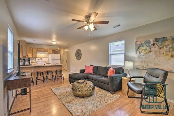 A modern living room with a sectional sofa, a coffee table, a dining area with bar stools, a kitchen, and a ceiling fan.