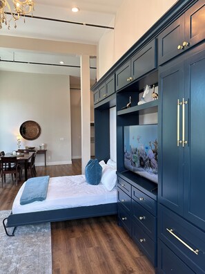 Een slaapkamer met een bed, een televisie, donkerblauwe kasten, een kroonluchter en een eethoek met een tafel en stoelen.