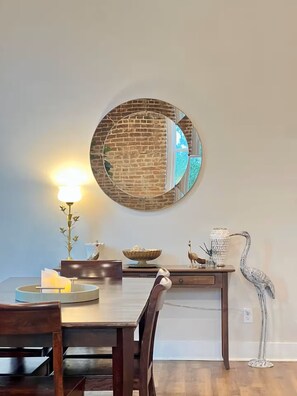 Een eethoek met een ronde spiegel, een tafel met stoelen, een lamp, decoratieve objecten en een hoge sculptuur.