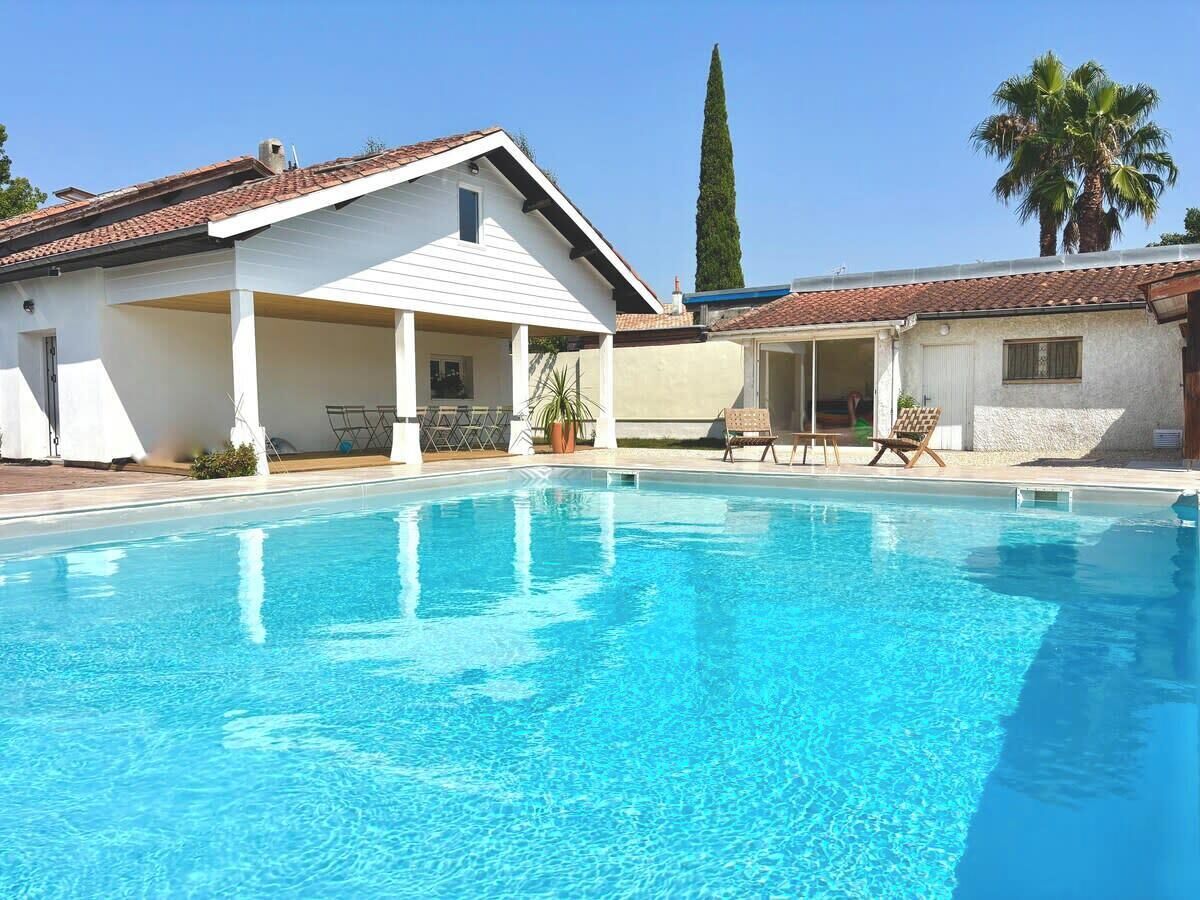 A swimming pool with a covered area, a tall tree, and a palm tree in the background.