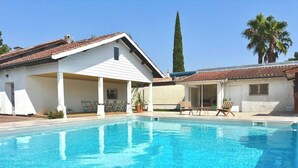 Una piscina con zona cubierta, un árbol alto y palmeras al fondo.