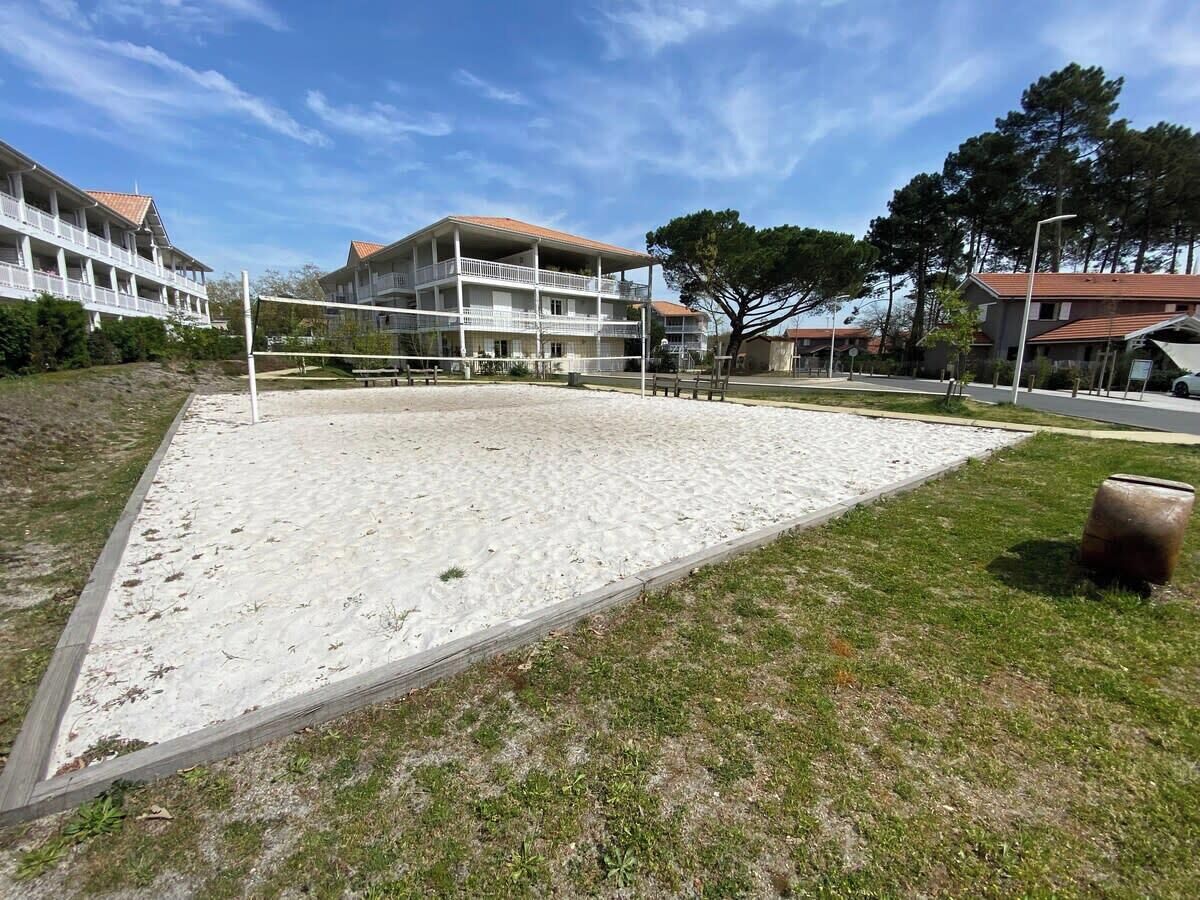 A sandy volleyball court with a wooden fence, surrounded by grass and trees, with buildings in the background.
