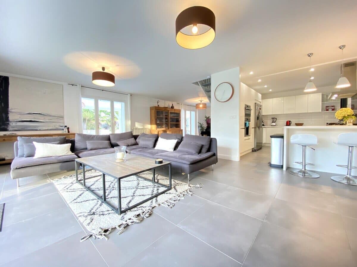 Een moderne woonkamer met een hoekbank, een salontafel en een keukenhoek met witte kasten en barstoelen.