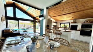Un coin repas avec une table dressée pour quatre personnes, une cuisine avec des meubles blancs et une vue dégagée sur l’extérieur grâce à de grandes fenêtres.