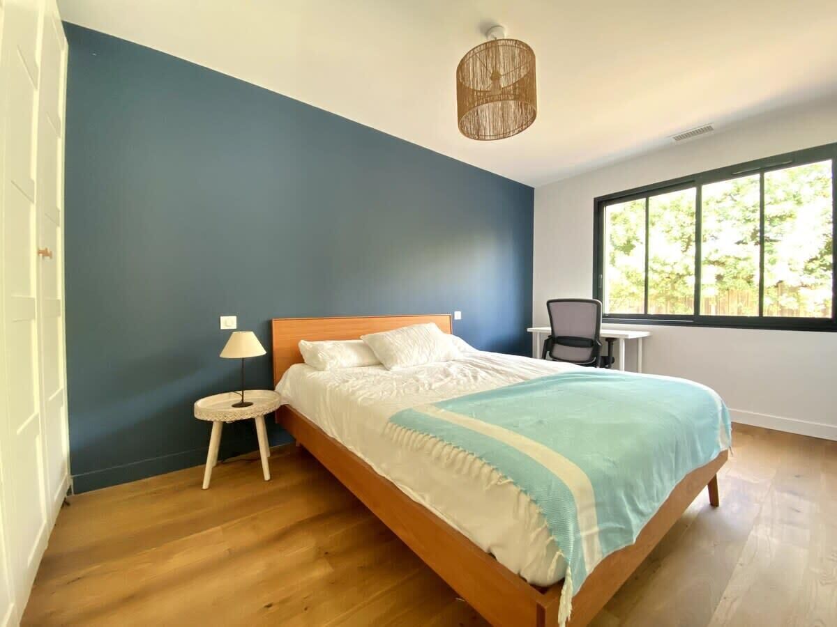 Un dormitorio con cama, mesita de noche, lámpara, silla y ventana con vistas a la vegetación.