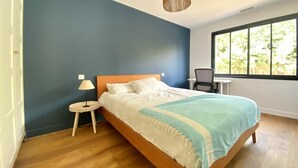 Ein Schlafzimmer mit Bett, Nachttisch, Lampe, Stuhl und einem Fenster mit Blick auf Grünflächen.