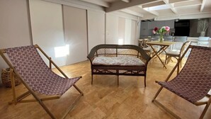 Una habitación con techo alto, un sofá de mimbre, dos sillones, una mesa de comedor con sillas y una zona de cocina al fondo.