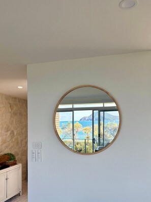 Ein Zimmer mit einem großen, runden Spiegel an der Wand, einem weißen Schrank mit einem Korb und Blick auf das Meer und die Berge durch ein Fenster.