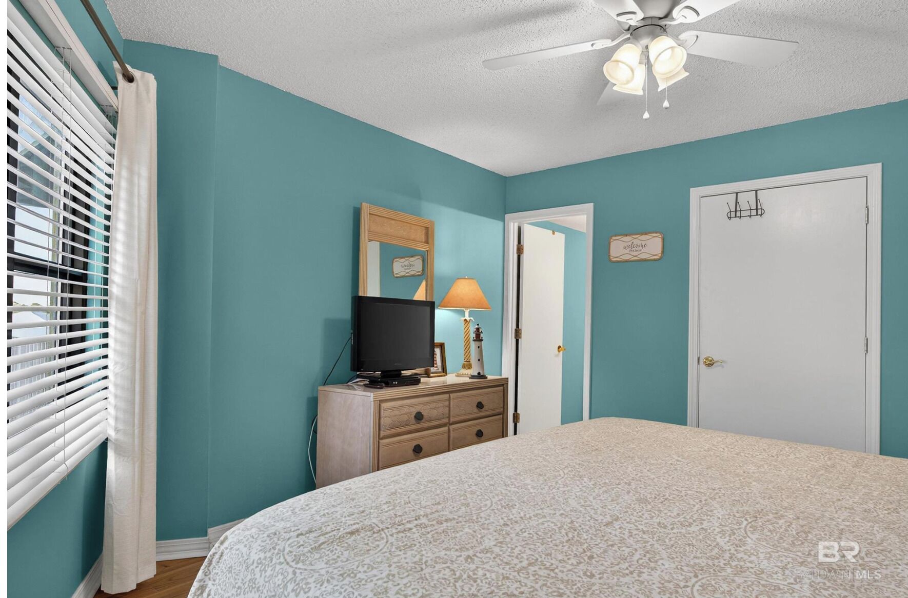 Une chambre avec des murs de couleur turquoise, un lit recouvert d’une couvre-lit à motifs, une commode sur laquelle est posée une télévision, un ventilateur de plafond et une porte ornée d’une pancarte « Bienvenue ».