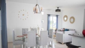 Ein modernes Wohnzimmer mit Essbereich, weißen Möbeln, einer Chandelüster und dekorativen Wandspiegeln.