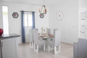 A dining area with a glass table, white chairs, a chandelier, and a window with curtains.