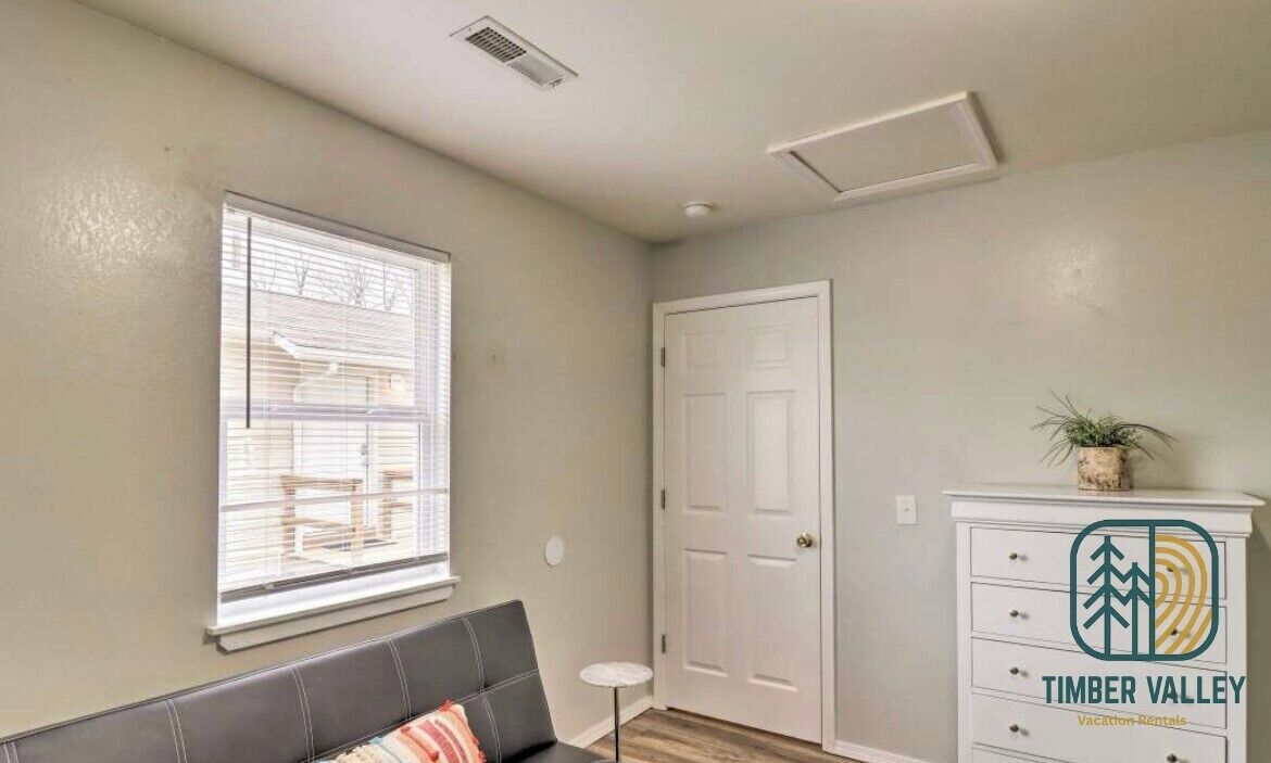 A room with a window, a door, a small table, a sofa, and a dresser with a logo.