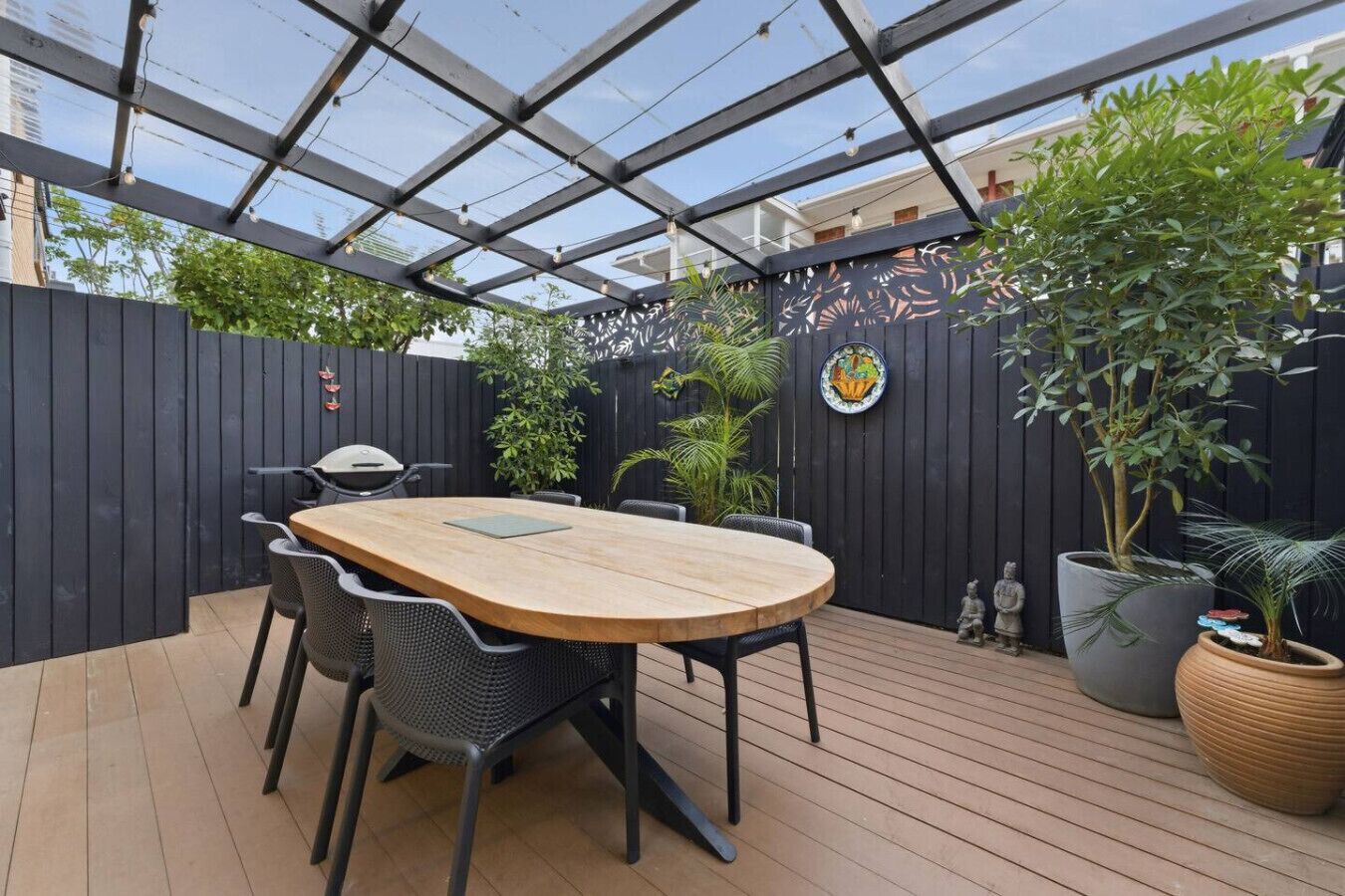 A covered outdoor patio with a wooden table and chairs, surrounded by potted plants and a decorative fence.