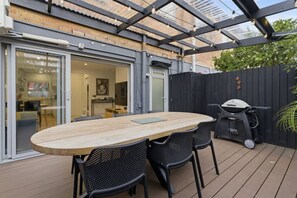 Eine Terrasse im Freien mit Holztisch und -stühlen, einem Grill und einem überdachten Bereich mit Blick ins Innere.