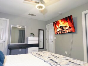 Ein Schlafzimmer mit Bett, Kommode, Spiegel, Deckenventilator und Fernseher, der ein Feuer zeigt.