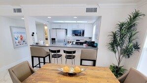 Uma área de refeições com uma mesa de madeira, cadeiras e um banco. Uma cozinha com eletrodomésticos modernos pode ser vista ao fundo.