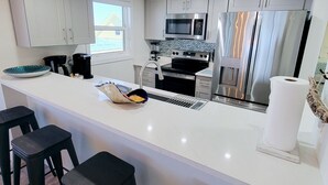 A modern kitchen with a central island, bar stools, stainless steel appliances, and a window providing natural light. - BEACH FRONT w/ ROOFTOP DECK! - {Aruba 6} Newly Renovated 2 BR / 2 BA - Walk to Times Square! (Fort Myers Beach)