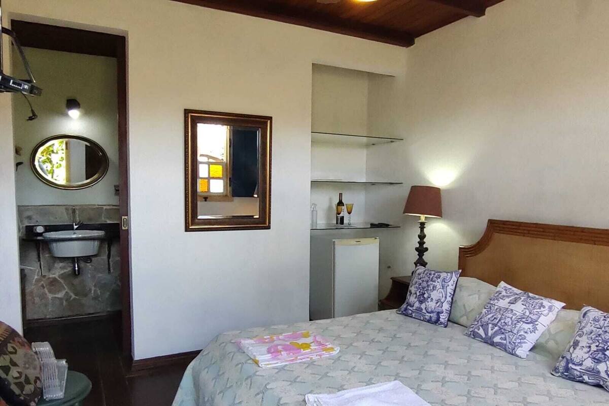 Une chambre à coucher comprenant un lit, une lampe de chevet, un miroir, une petite table, ainsi qu’une salle de bain avec un lavabo et un miroir.