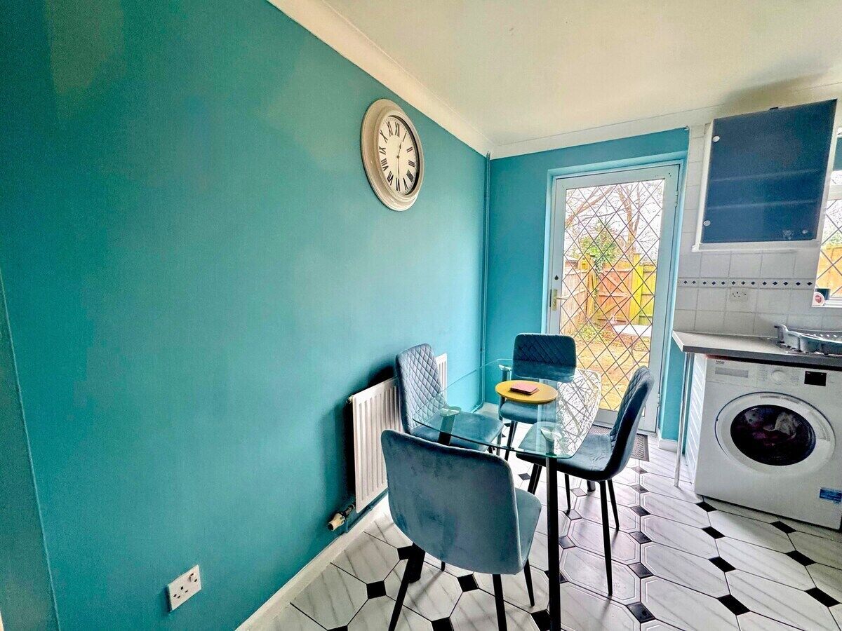 A compact kitchen with a washing machine, a dining table with chairs, and a clock on the wall.
