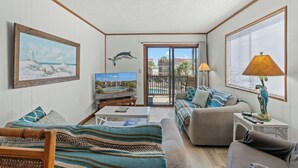 A living room with a sofa, a television, a table, a lamp, and a view of a pool and buildings through a glass door. - Inlet Point Villas 11D - Pool View (North Myrtle Beach)