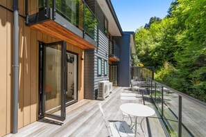 A modern house with a deck, outdoor furniture, and a glass railing overlooking a forested area. - Queenstown Forest Lodge, 4BR Retreat with Spa Pool, Views, Games & Family Comfort (Queenstown)