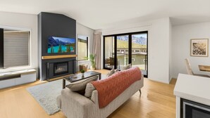 A modern living room with a fireplace, a sofa, a coffee table, and a TV. - Forest Creek Lodge– Mountain Views & Spa Pool (Queenstown)