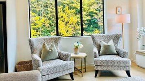 A room with two patterned armchairs, a small round table with a vase, and a large window with a view of yellow foliage. - Forest Creek Lodge– Mountain Views & Spa Pool (Queenstown)