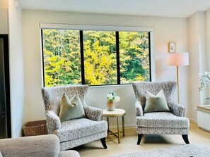 Ein Zimmer mit zwei gemusterten Sesseln, einem kleinen runden Tisch mit einer Vase und einem großen Fenster mit Blick auf gelbes Laub.