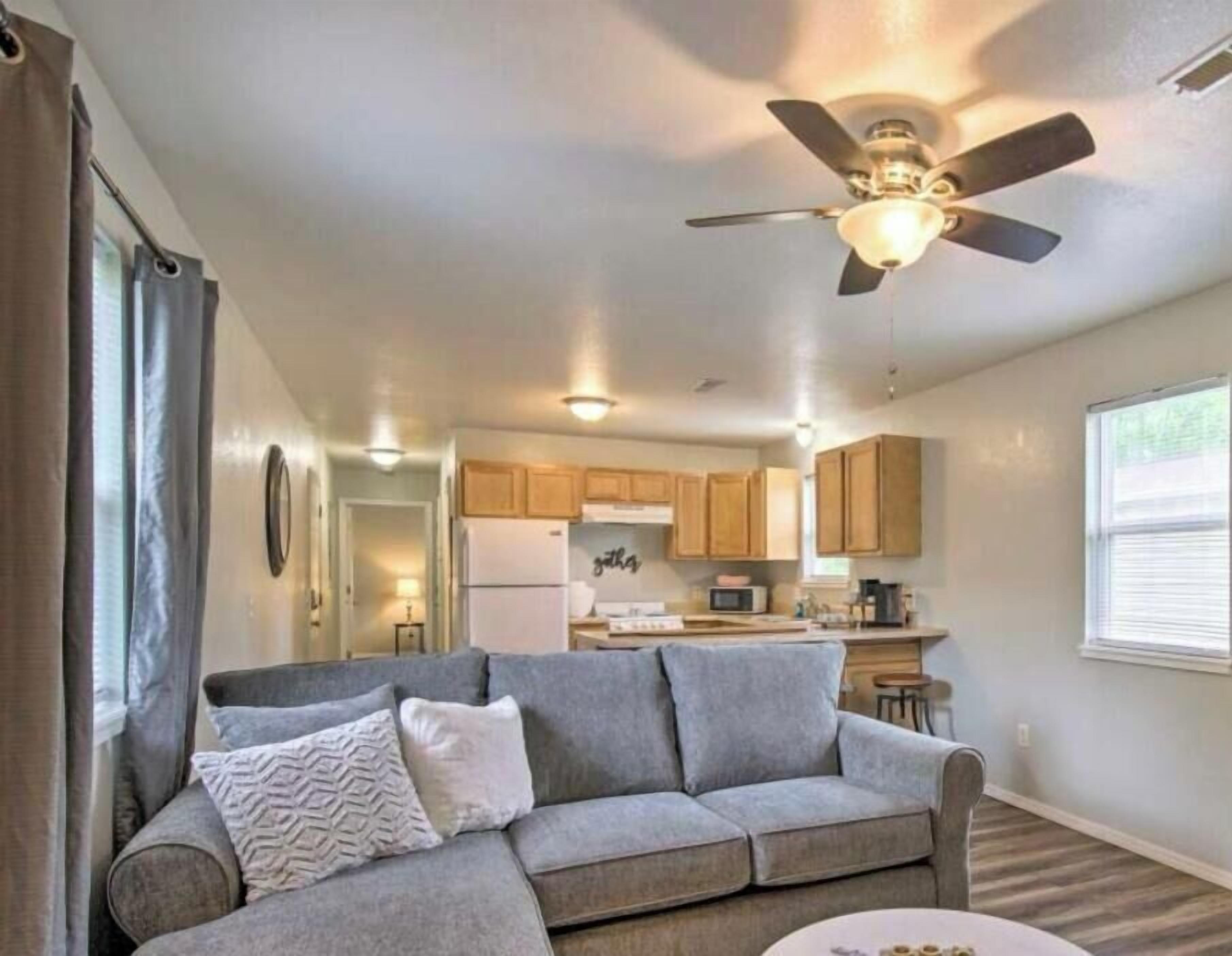Un salón con un sofá gris, un ventilador de techo y una cocina al fondo.