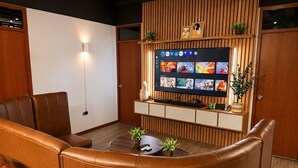 A living room with a leather sofa, a coffee table with plants, a flat-screen TV mounted on a wooden wall, and a wooden cabinet below.