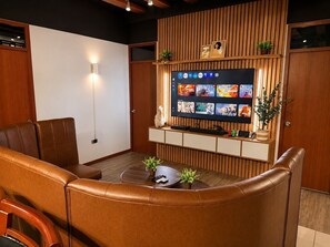 A living room with a leather sofa, a coffee table with plants, a flat-screen TV mounted on a wooden wall, and a wooden cabinet below.