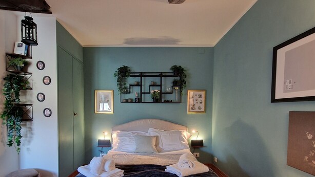 Quarto com cama, abajures, prateleira na parede com plantas, tapete e um banquinho.