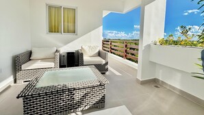 A balcony with wicker furniture, a glass table, and a view of greenery and a clear sky. - Perfect 2-bedroom building with AC in welcoming Punta Cana (Punta Cana)
