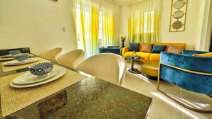 A living room with a yellow sofa, a dining table with bowls, and a painting on the wall. - Perfect 2-bedroom building with AC in welcoming Punta Cana (Punta Cana)