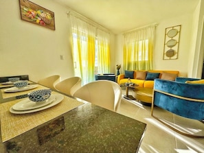 A living room with a yellow sofa, a dining table with bowls, and a painting on the wall. - Perfect 2-bedroom building with AC in welcoming Punta Cana (Punta Cana)