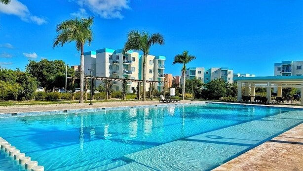 A swimming pool with clear blue water, surrounded by palm trees and modern buildings in the background. - Perfect 2-bedroom building with AC in welcoming Punta Cana (Punta Cana)