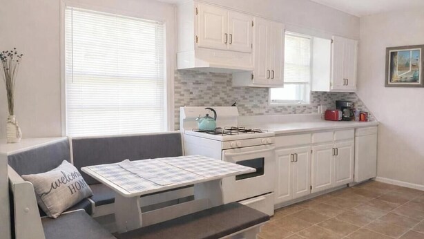 A compact kitchen with white cabinets, a gas stove, a small dining table with a bench, and a window with blinds. - Quintessential cottage walking distance to Oaklawn and minutes from downtown (Hot Springs)