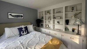 A bedroom with a bed, a headboard, a wall-mounted shelf unit, and a bedside table with a lamp.