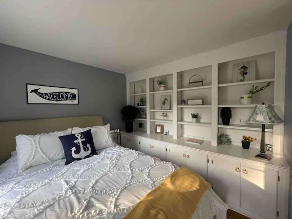 A bedroom with a bed, a headboard, a wall-mounted shelf unit, and a bedside table with a lamp.
