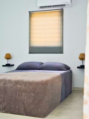 Un dormitorio con cama, dos mesitas de noche con lámparas, una ventana con persianas y un equipo de aire acondicionado en la pared.