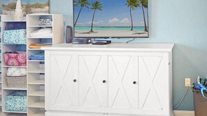 Quarto com televisão em um armário branco, uma estante com bolsas estampadas e um quadro de praia na parede.