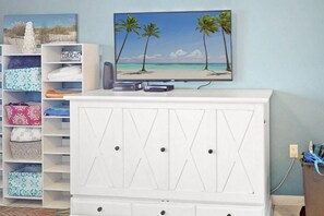 Quarto com televisão em um armário branco, uma estante com bolsas estampadas e um quadro de praia na parede.