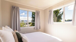 A bedroom with a bed, curtains, and a view of greenery and mountains. - Classic 3 Bedroom Holiday Home (Twizel)