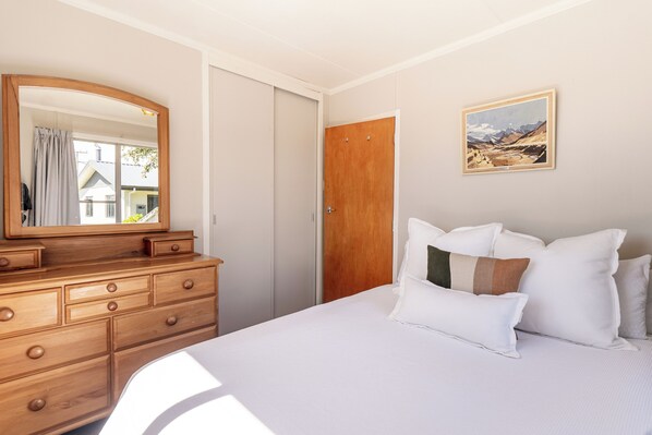 A bedroom with a wooden dresser, a bed with white bedding, a wooden door, and a framed landscape painting on the wall.