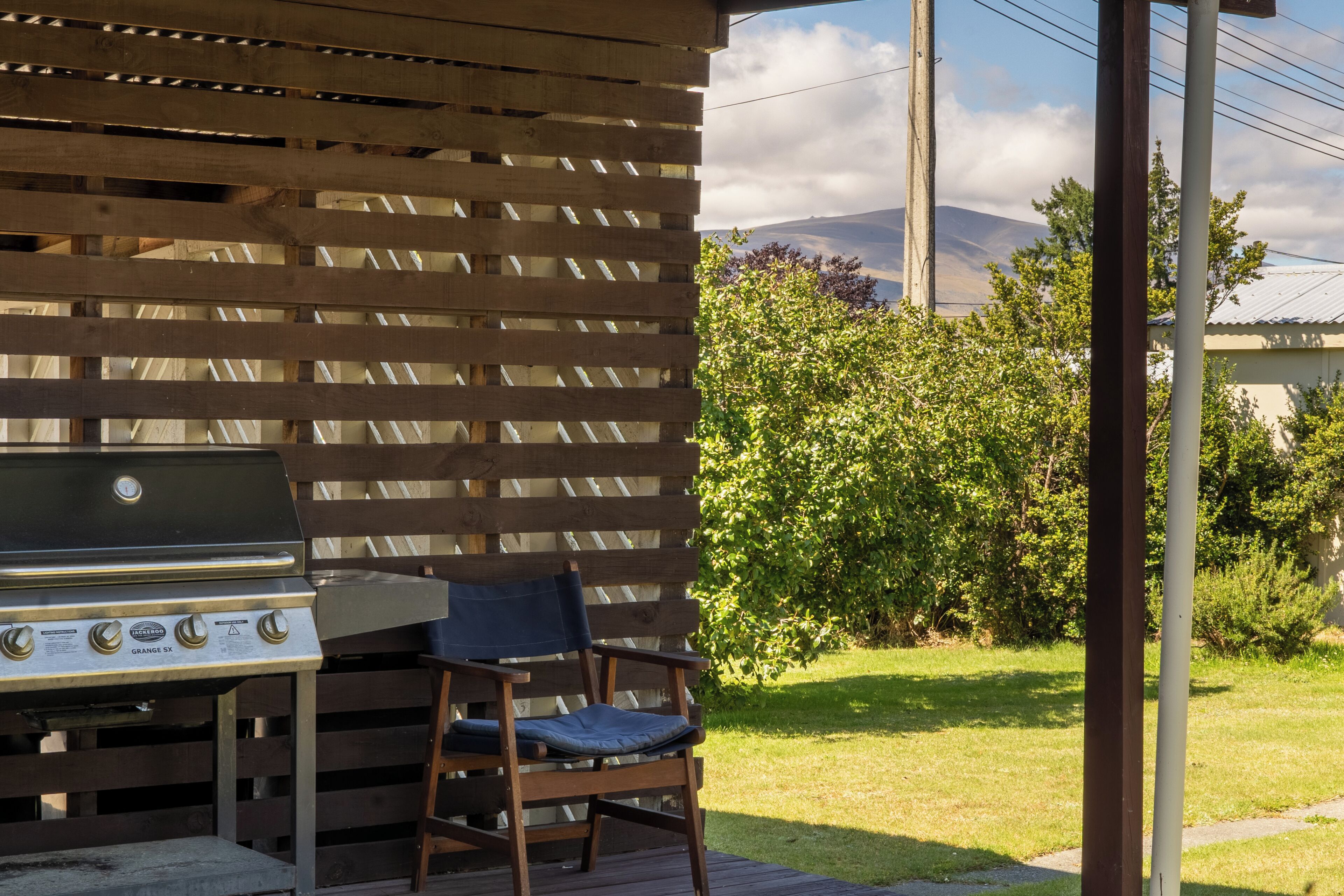 A barbecue grill with a wooden table and chair, set against a wooden fence with a view of a grassy area and trees.